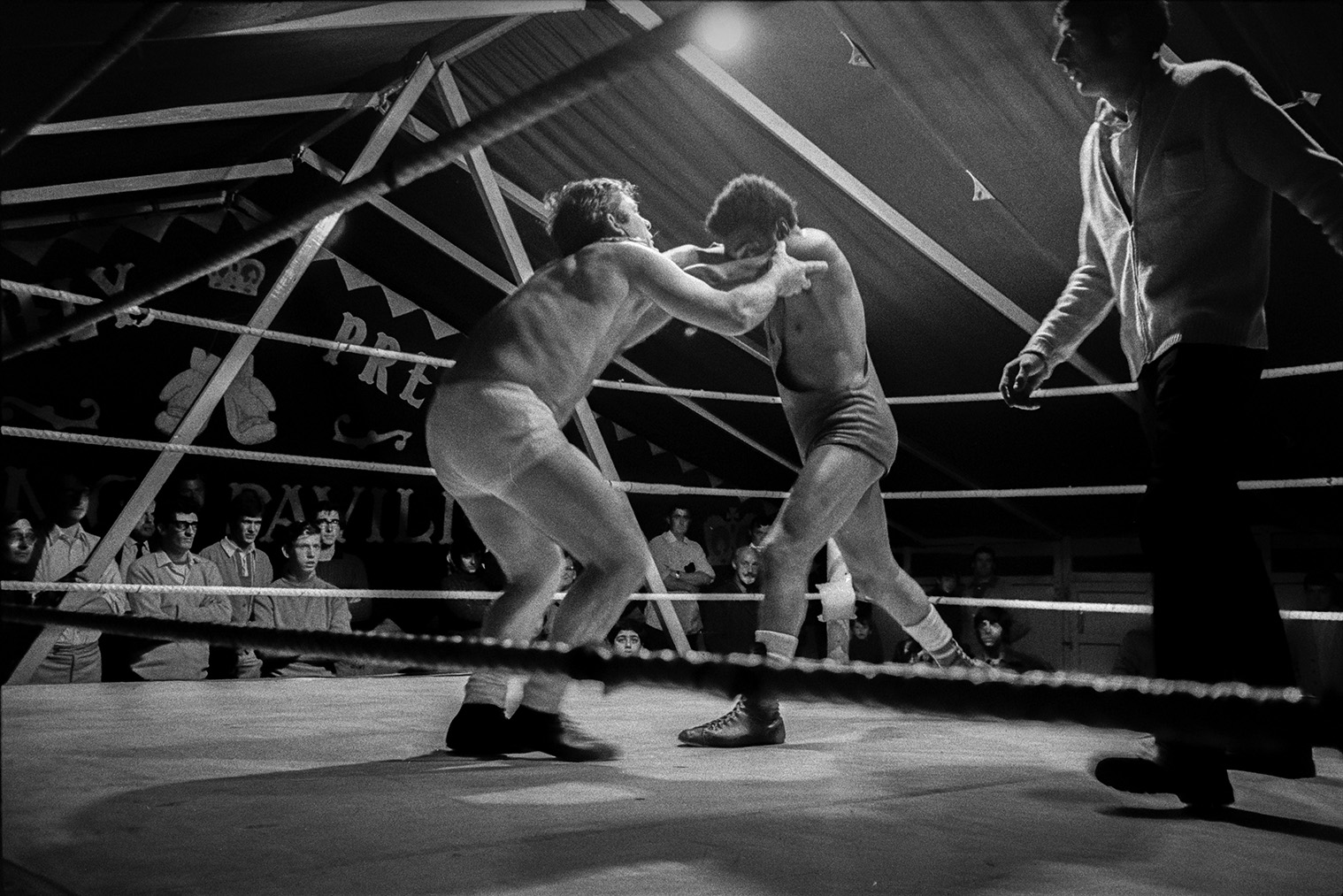 A wrestling match in a tent at Barnstaple Fair. Spectators are watching around the ring and the referee is supervising the match.