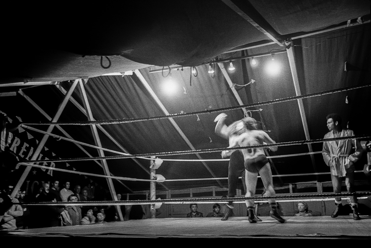 A wrestling match in a tent at Barnstaple Fair. Spectators are watching around the ring and another man is waiting to fight.