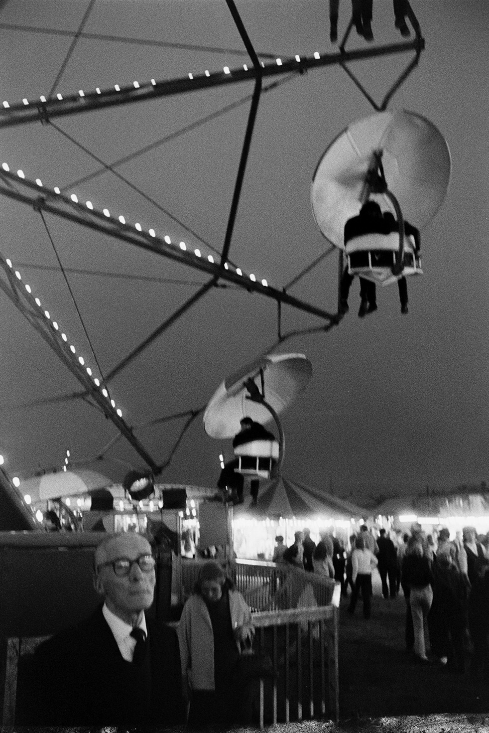 A man and woman stood by a big wheel fairground ride at Barnstaple Fair. Other people and fairground stalls can be seen in the background.