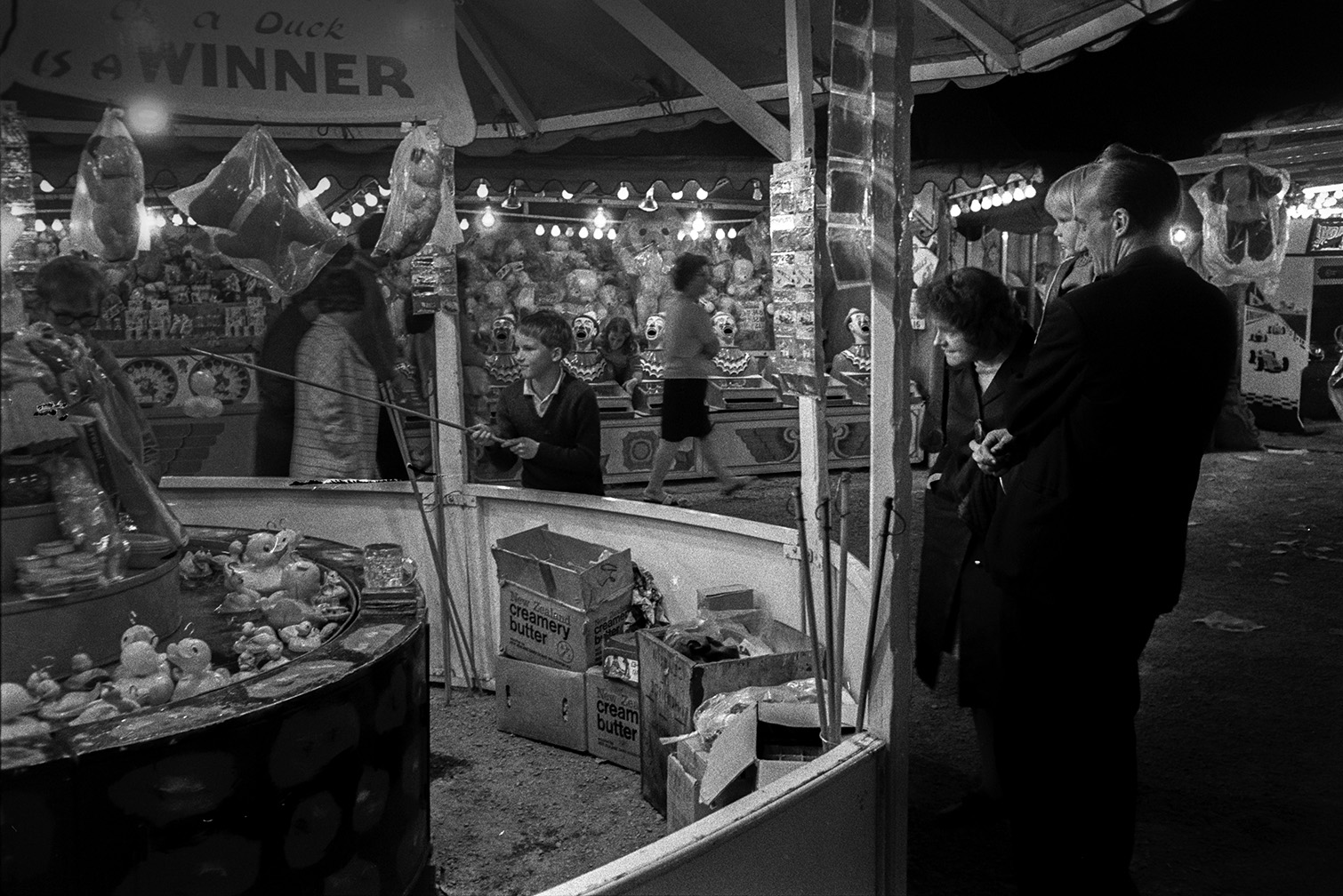 A boy playing the 'hook a duck' game at Barnstaple Fair. His family are watching. Various prizes, including soft toys, are hung up on the stall. Another fairground game can be seen in the background.