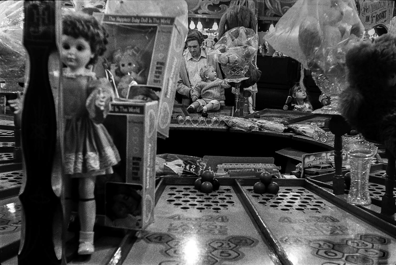 A man and children playing games at Bideford Fair. Various prizes are on display including  dolls.