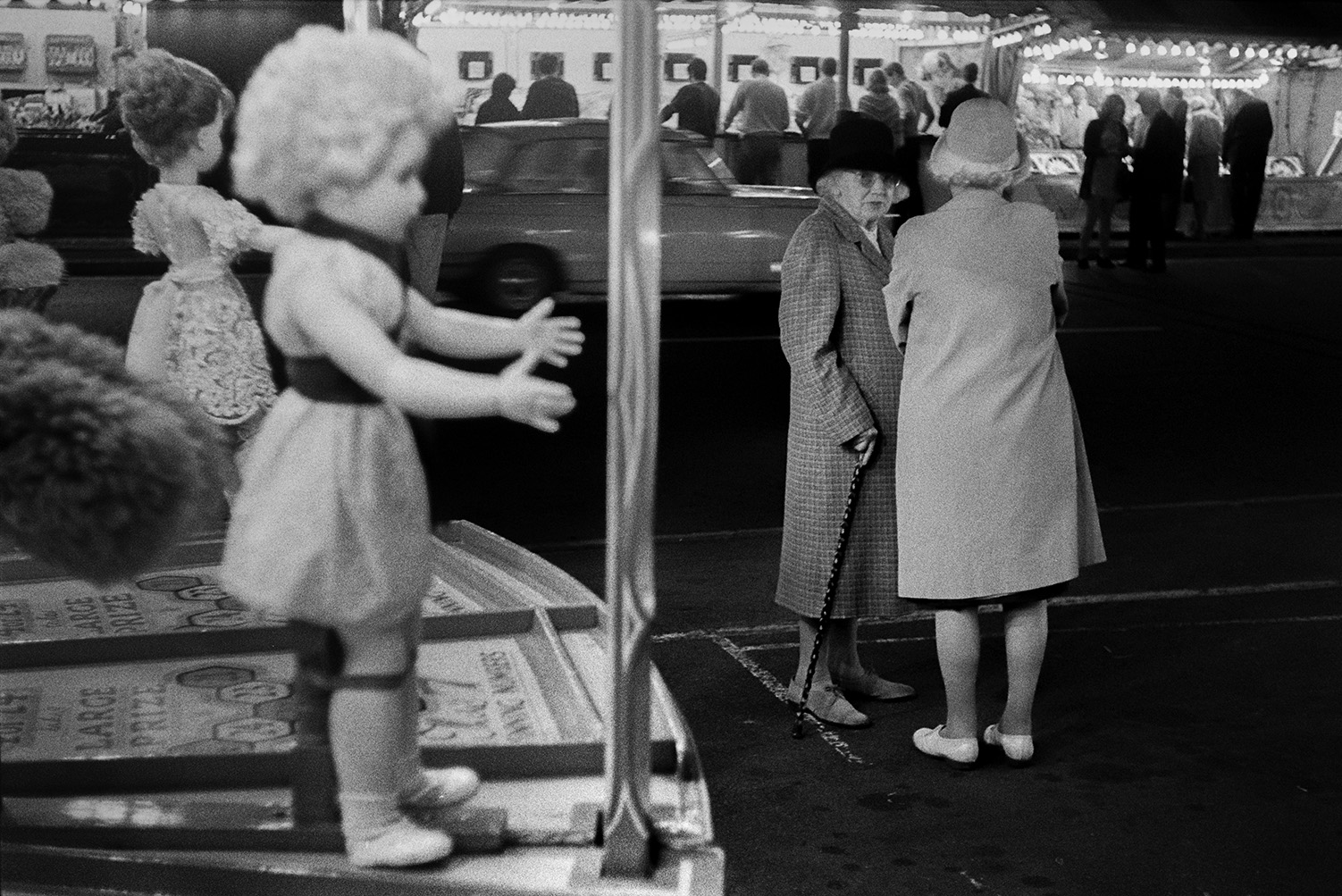 Two women talking at Bideford Fair by a fairground game with dolls. One of the women has a walking stick. Other people can be seen at fairground stalls in the background.