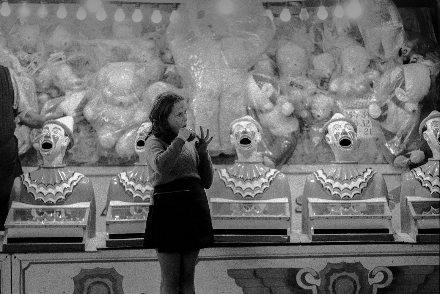 A woman eating a toffee apple stood by a clown game at Barnstaple fair. Soft toy prizes can be seen behind the game.
