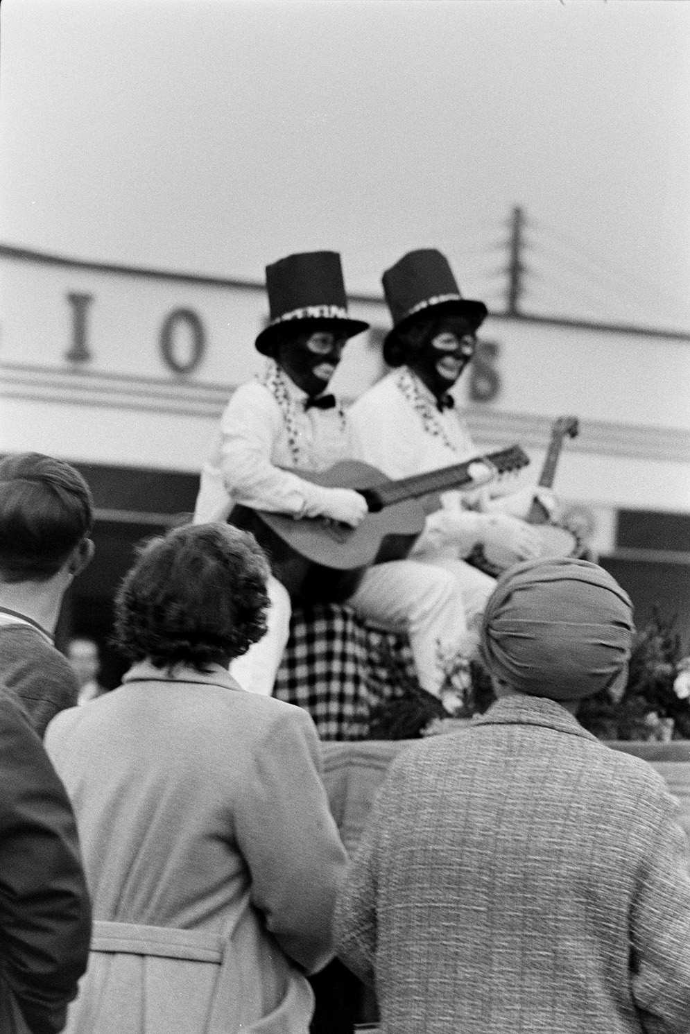 People watching Bideford carnival procession. Two people on one of the carnival floats are dressed as Black and White Minstrels, and playing a guitar and banjo.