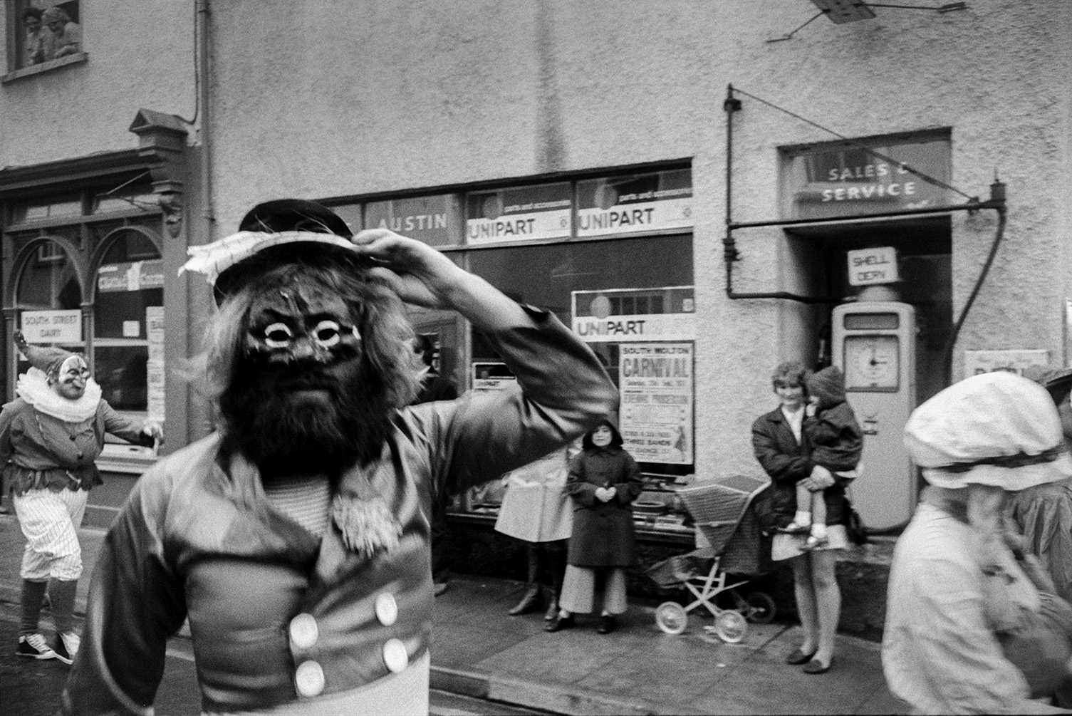 Spectators outside a shop front watching people in masks and costumes in South Molton carnival parade. A woman holding a child is stood by a pram and petrol pump, watching the parade.