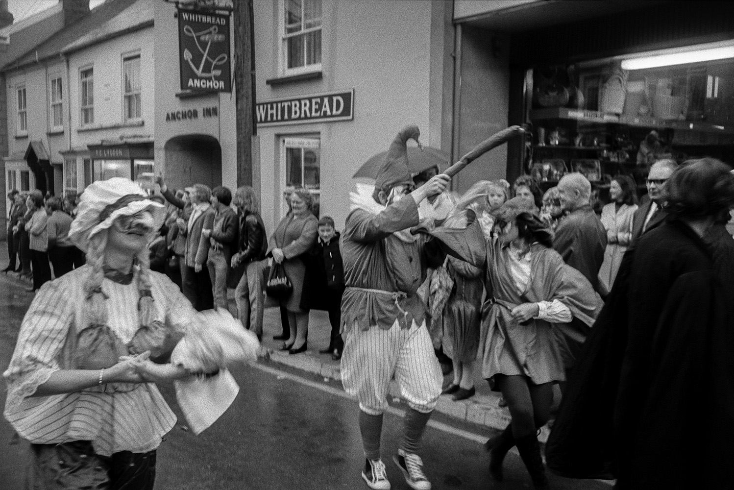 Spectators outside the Anchor Inn watching people in costumes and masks dancing through South Street in the South Molton carnival parade. The Inn is now Hungs Chinese Restaurant. The shop next door, Kingdon's Hardware Shop, is now The Factory Shop.