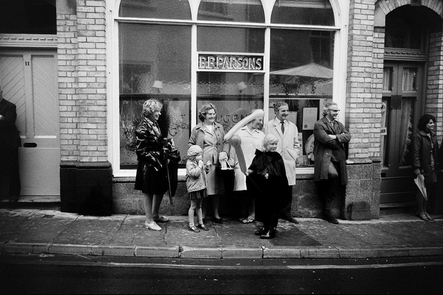 Spectators stood outside B B Parsons shopfront watching the South Molton carnival parade. One of the children is holding a balloon. The shop later became a hairdressers and then a chiropractors.