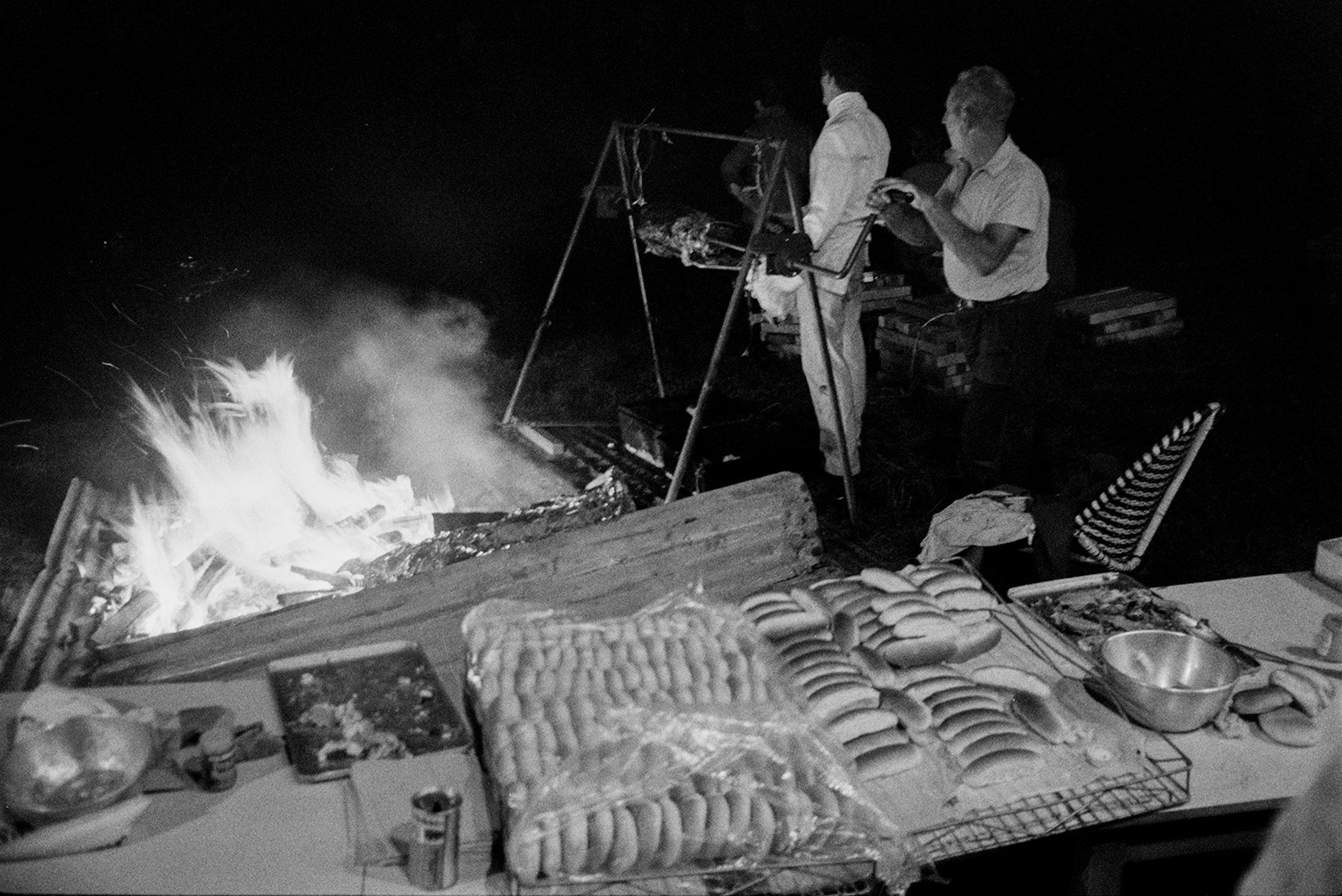 A man turning a spit roast by an open air fire at Torrington Water Carnival. A table with bread rolls and food is visible in the foreground.