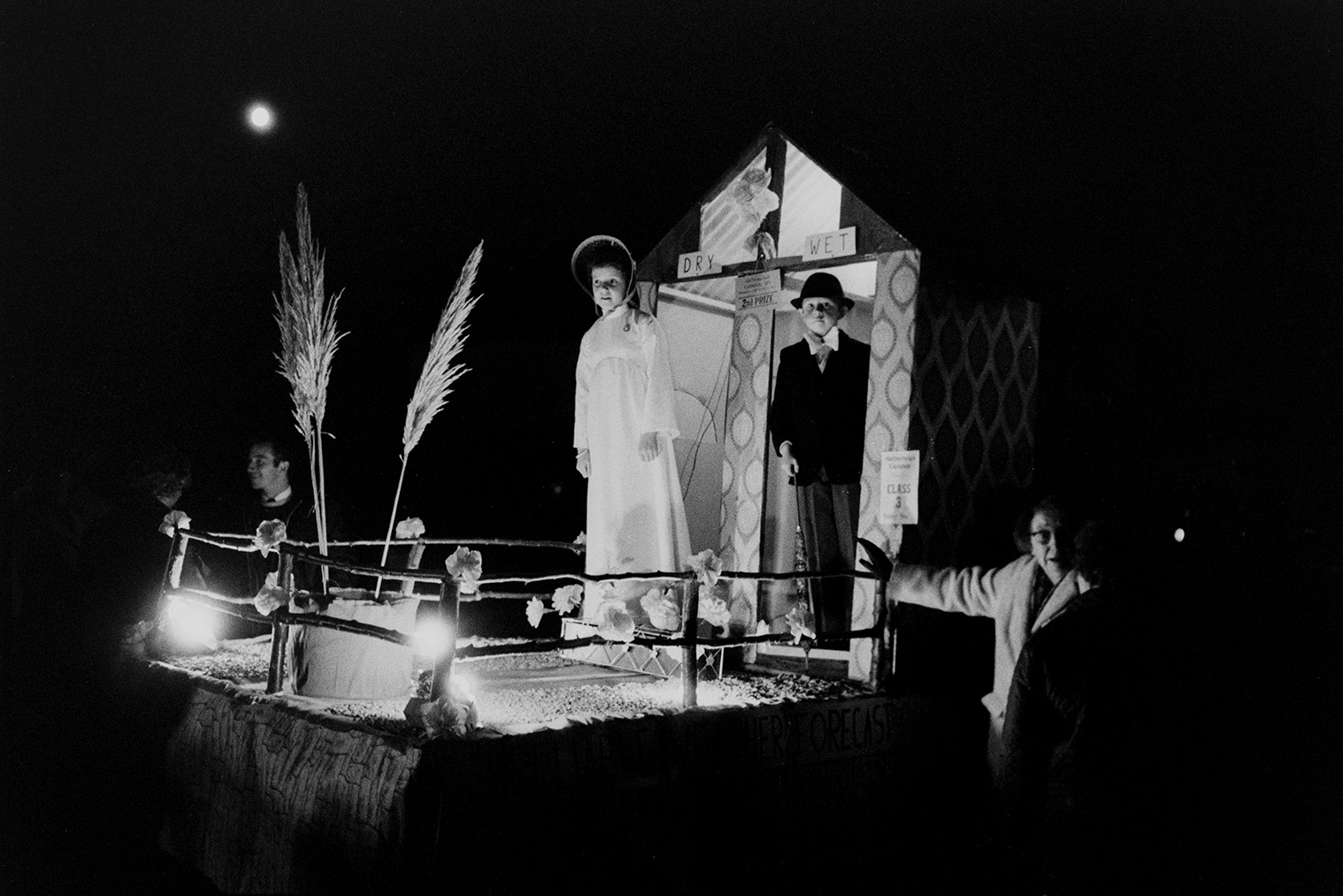 A girl and boy on a weather vane themed carnival float at Hatherleigh Carnival, at night. A woman is walking beside the float.