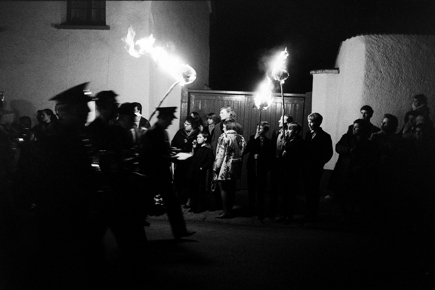 Spectators holding burning torches watching band members parading in Hatherleigh Carnival at night.