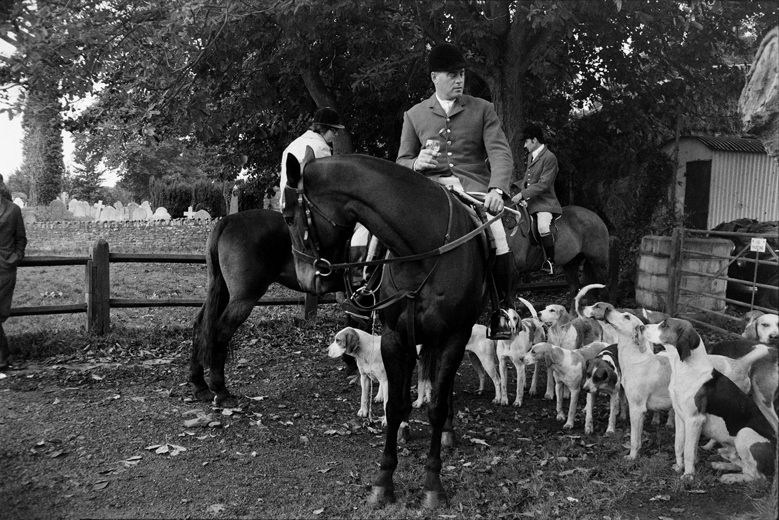 Tavistock Staghound hunt meet at a farm in Lapford before setting off on a hunt. Mounted huntsmen are drinking their stirrup cups and hounds are gathered around. Gravestones in a churchyard are visible in the background.