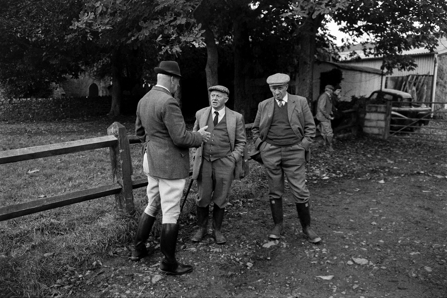 Three men talking at the Tavistock Staghound hunt meet at a farm in Lapford. They are stood by a wooden fence under trees. Farm buildings and gravestones in a churchyard can be seen in the background.