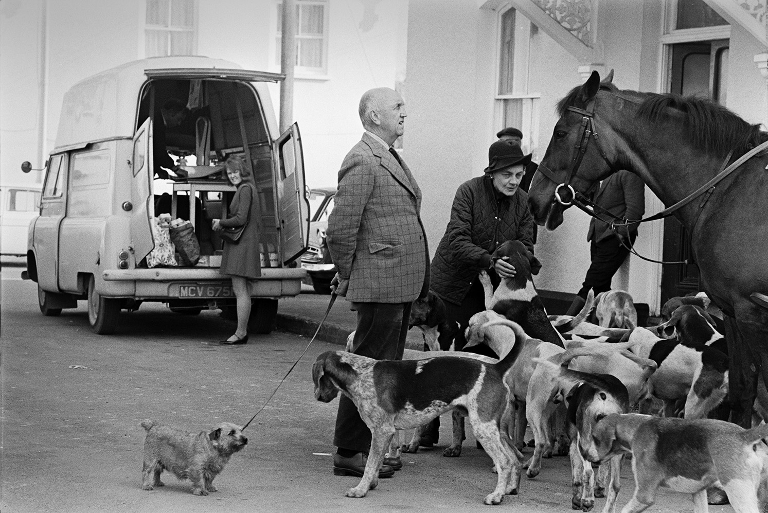 Tiverton Foxhounds hunt meet in village square at Witheridge. Spectators are greeting the hounds and a mounted huntsman. The man has a dog with him. A woman is getting items out of a van parked in the square in the background.