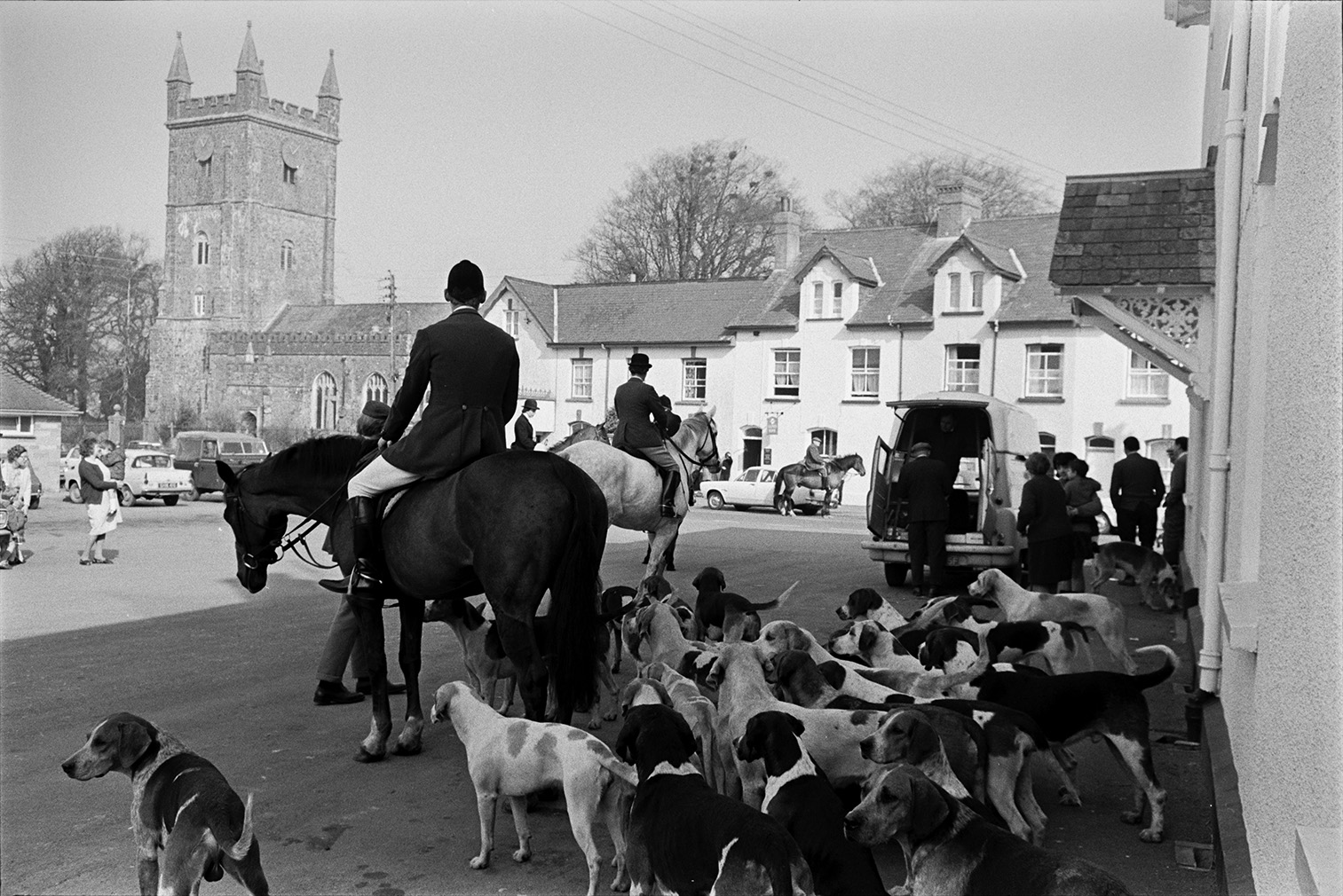 Tiverton Foxhounds hunt meet in the village square at Witheridge. Spectators have gathered to watch the mounted huntsmen and hounds. The Church and house can be seen in the background.