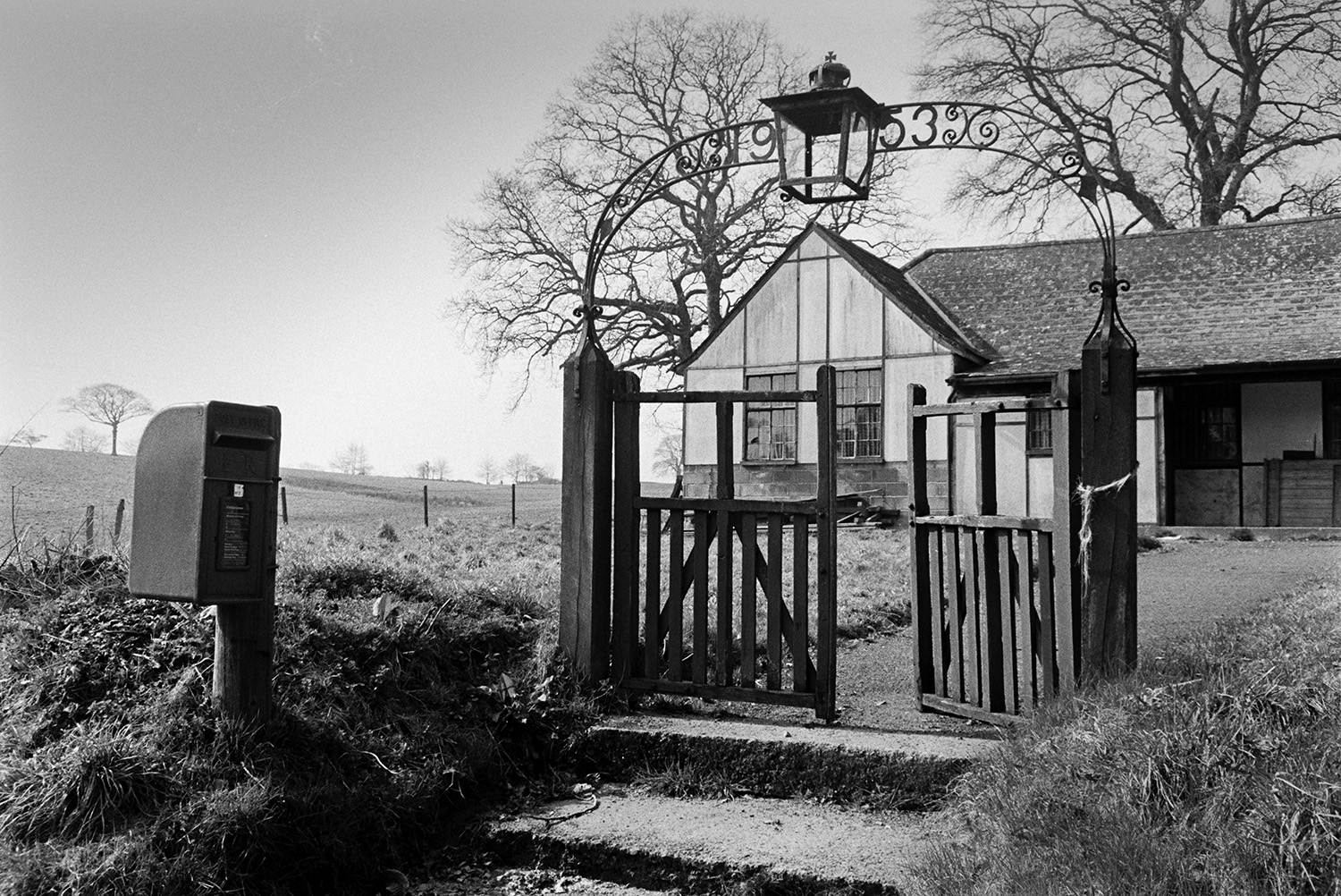 A bungalow with wooden gate, lamp and post box at its entrance, possibly in Witheridge. The date 1953 is worked into to design of the ironwork above the gate.
