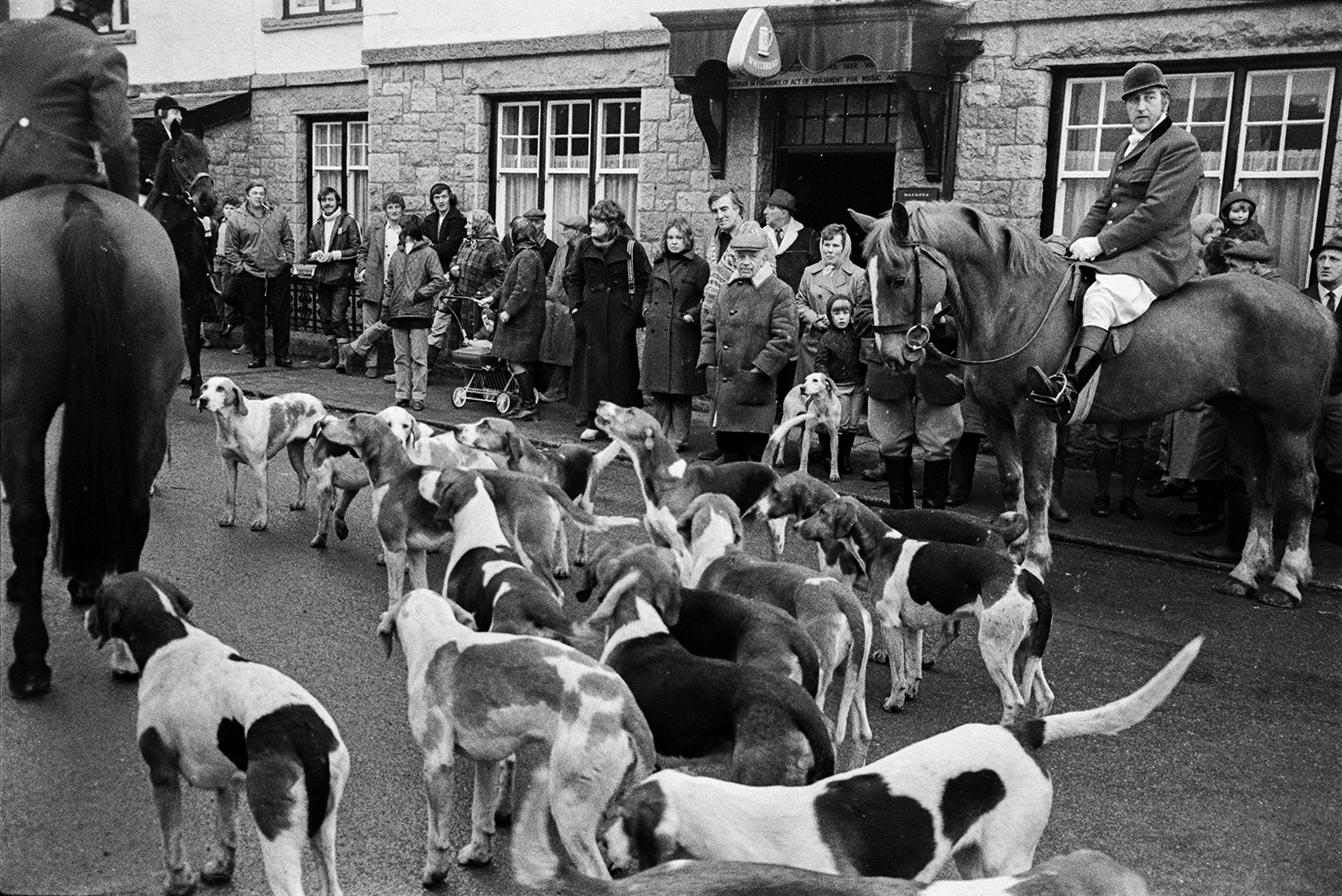 Spectators watching a Torrington Farmers Hunt meet outside a pub in Winkleigh. Mounted huntsmen and hounds are visible.