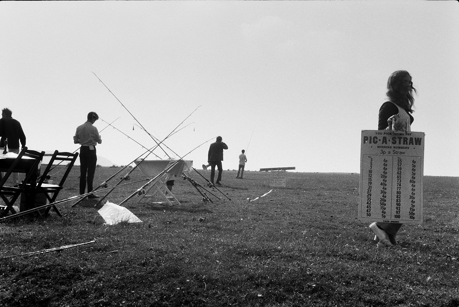Appledore Angling Contest at Westward Ho! Fishing rods are stacked against a table with competitors stood around. A woman in the foreground is advertising a straw game.