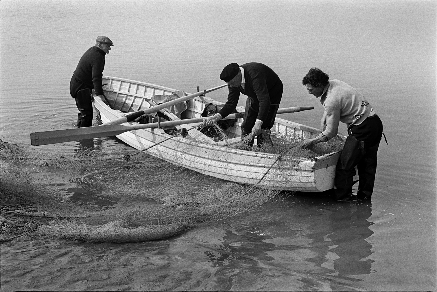 Two men hauling a fishing net into rowing boat, possibly at Appledore. Two oars are resting across the boat and another man is stood in the water and leaning on the rowing boat.