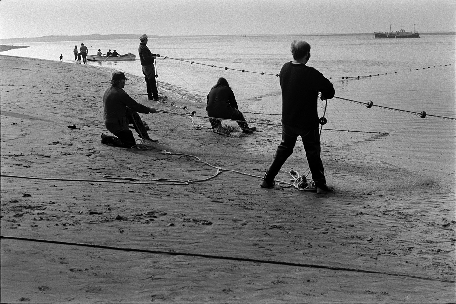 Men netting salmon on a beach front, possibly at Appledore. A ship can be seen on the water and people in a rowing boat are visible in the background.