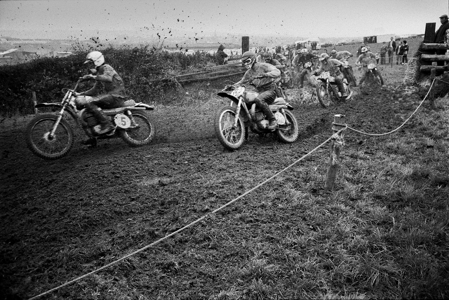 Competitors racing along a muddy course at Torrington motorbike scramble. Mud is flying up from the motorbike wheels. Spectators are watching in the background.