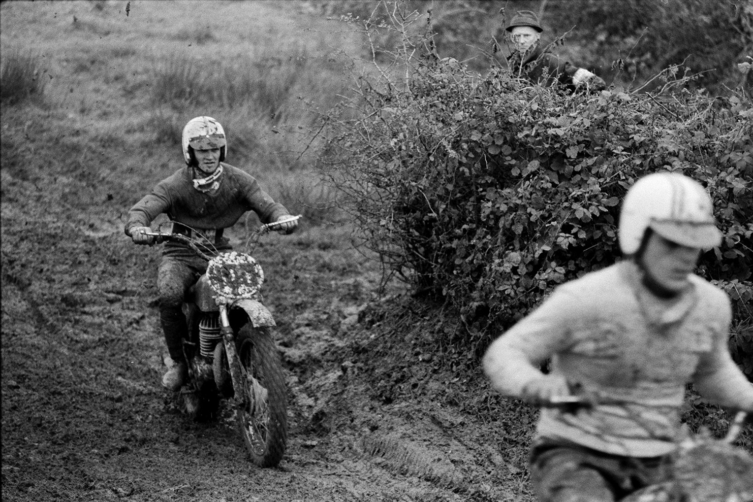 Competitors racing along a muddy course at Torrington motorbike scramble. A man is watching them over a hedge.