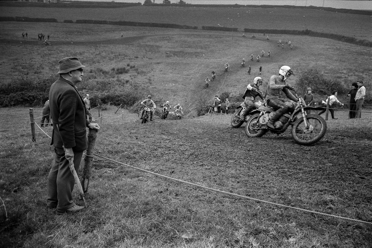 Spectators, standing behind rope barriers, watching competitors racing along a muddy course at Torrington motorbike scramble