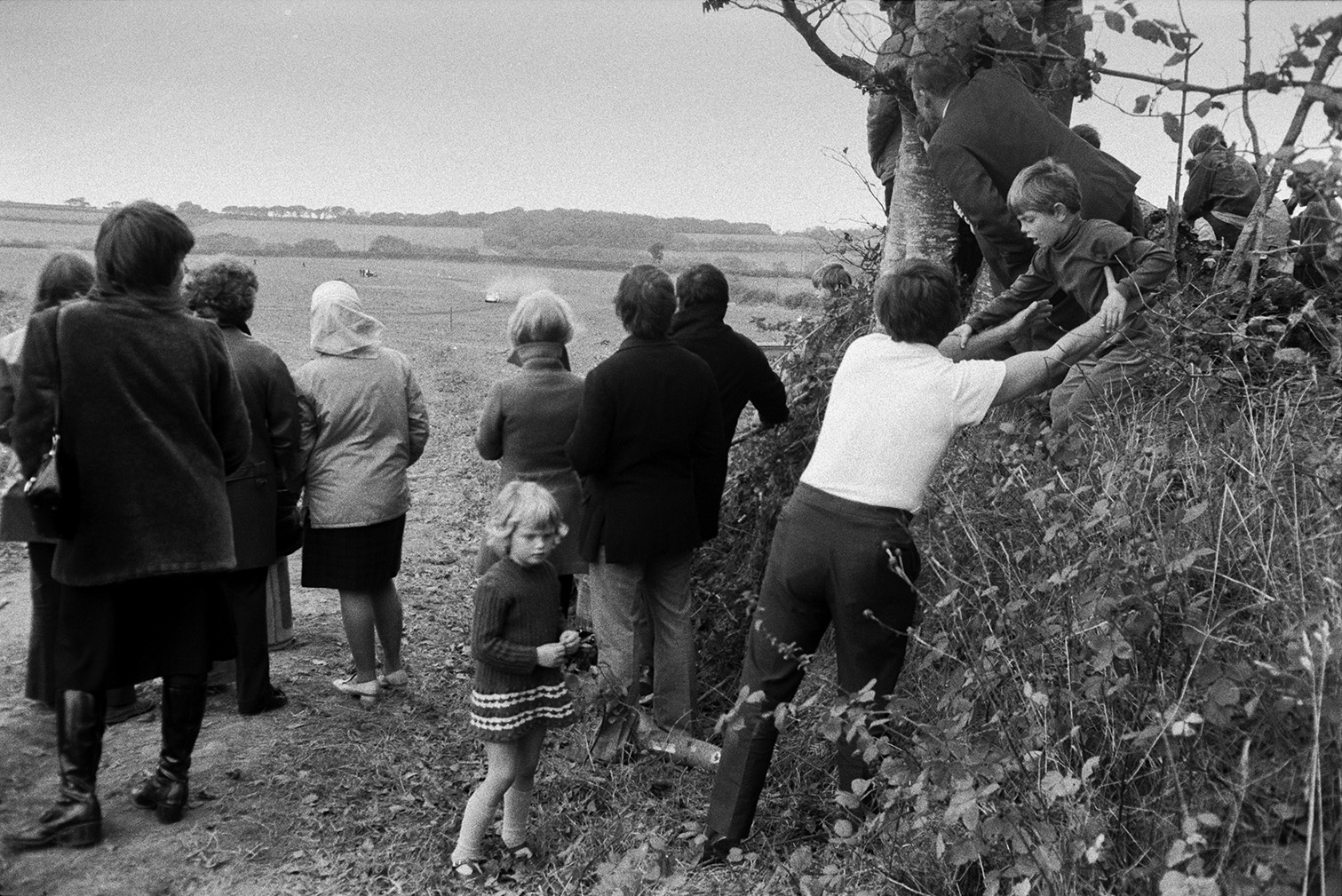Spectators watching banger car racing at Sugworthy. A man is helping a child down from a hedge where he has been stood to get a better view. The bangers cars can be seen on a track in the distance.