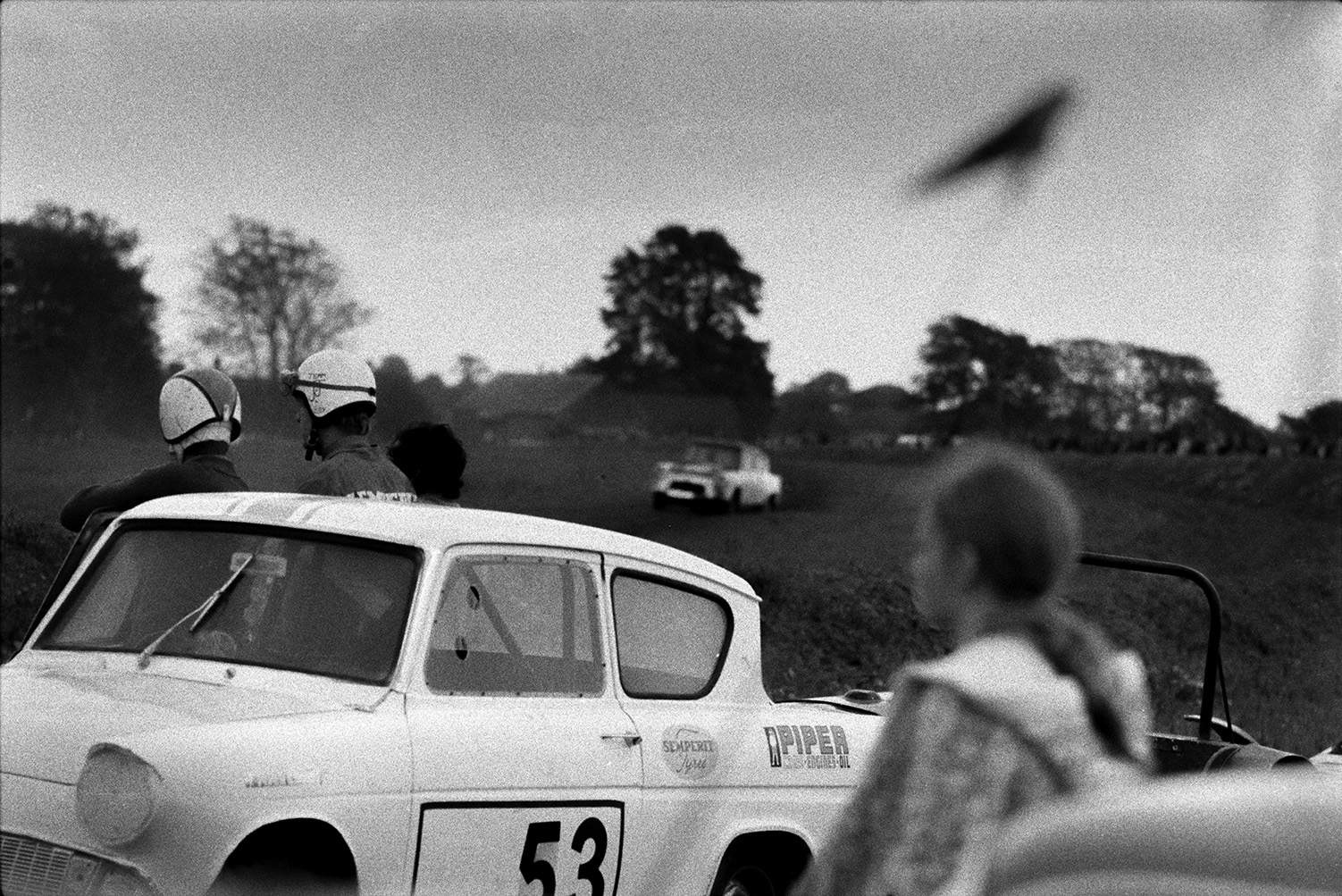 Two competitors stood by a banger car before racing it at Sugworthy. Another car can be seen racing in the background and a girl is watching in the foreground.