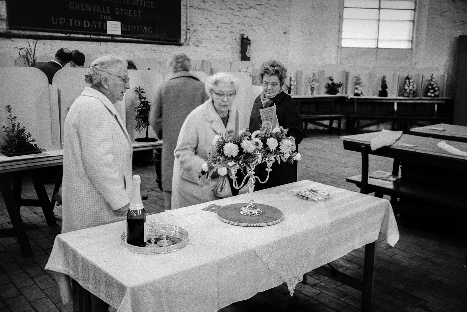 Three women looking at a flower arrangement in a candelabra at a flower show in Bideford Pannier Market. A tray with a bottle of champagne and glasses is next to the flower display.