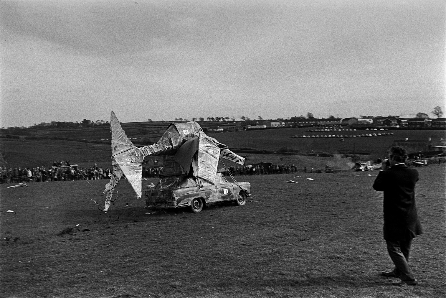 Spectators watching man made flying machines try to take off at a 'Man Powered Flight' competition in a field at Crowbeare. A man is either taking a photograph or filming a car with a fish model on top in the foreground.