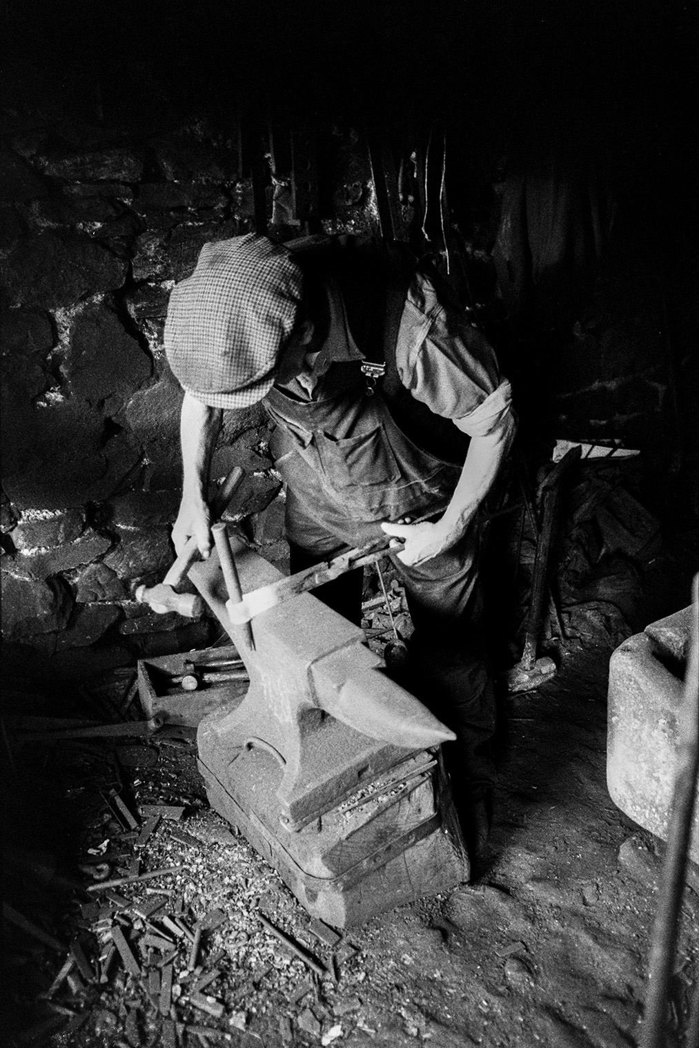 Mr Loosemore, a Blacksmith, working in his forge at Atherington to make hinges for wooden farm gates. He is beating a piece of heated metal over an anvil.