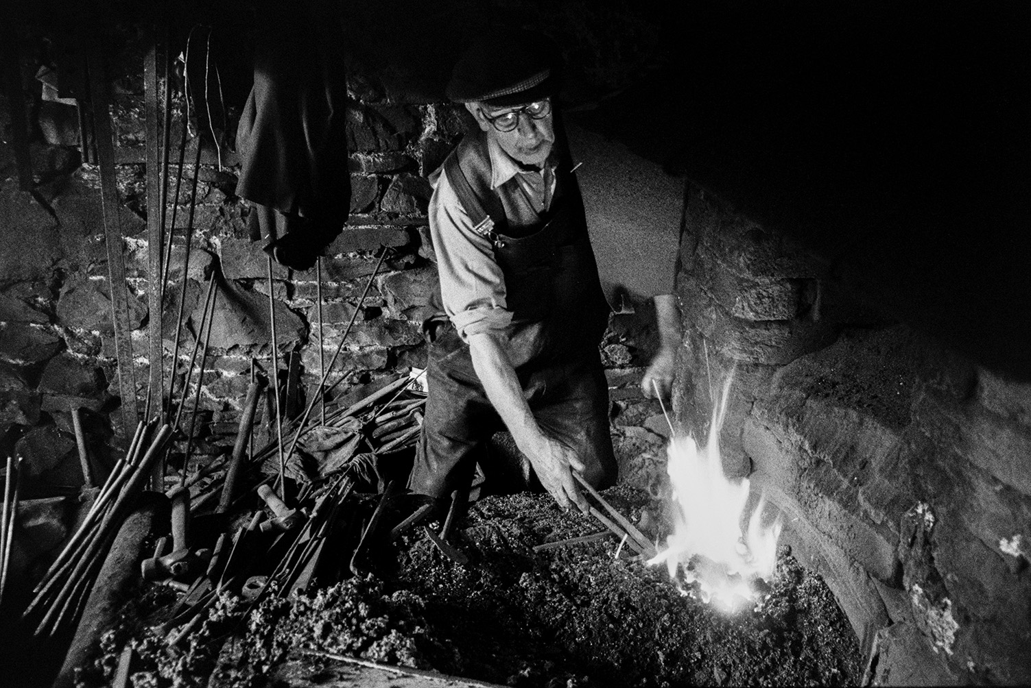 Mr Loosemore, a Blacksmith, working in his forge at Atherington to make hinges for wooden farm gates. He is holding the hinges using a pair of tongs and heating them up in a fire.