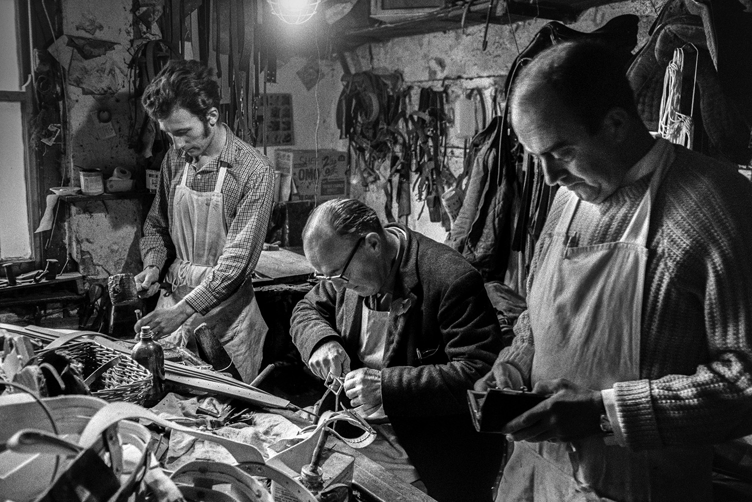 Three men including Walter Johns, sat at the workbench, Derek Johns, on the right and Mr Bowden, on the left, working on leather goods in their saddlery workshop in Bideford. Various ropes and tools can be seen in the workshop.