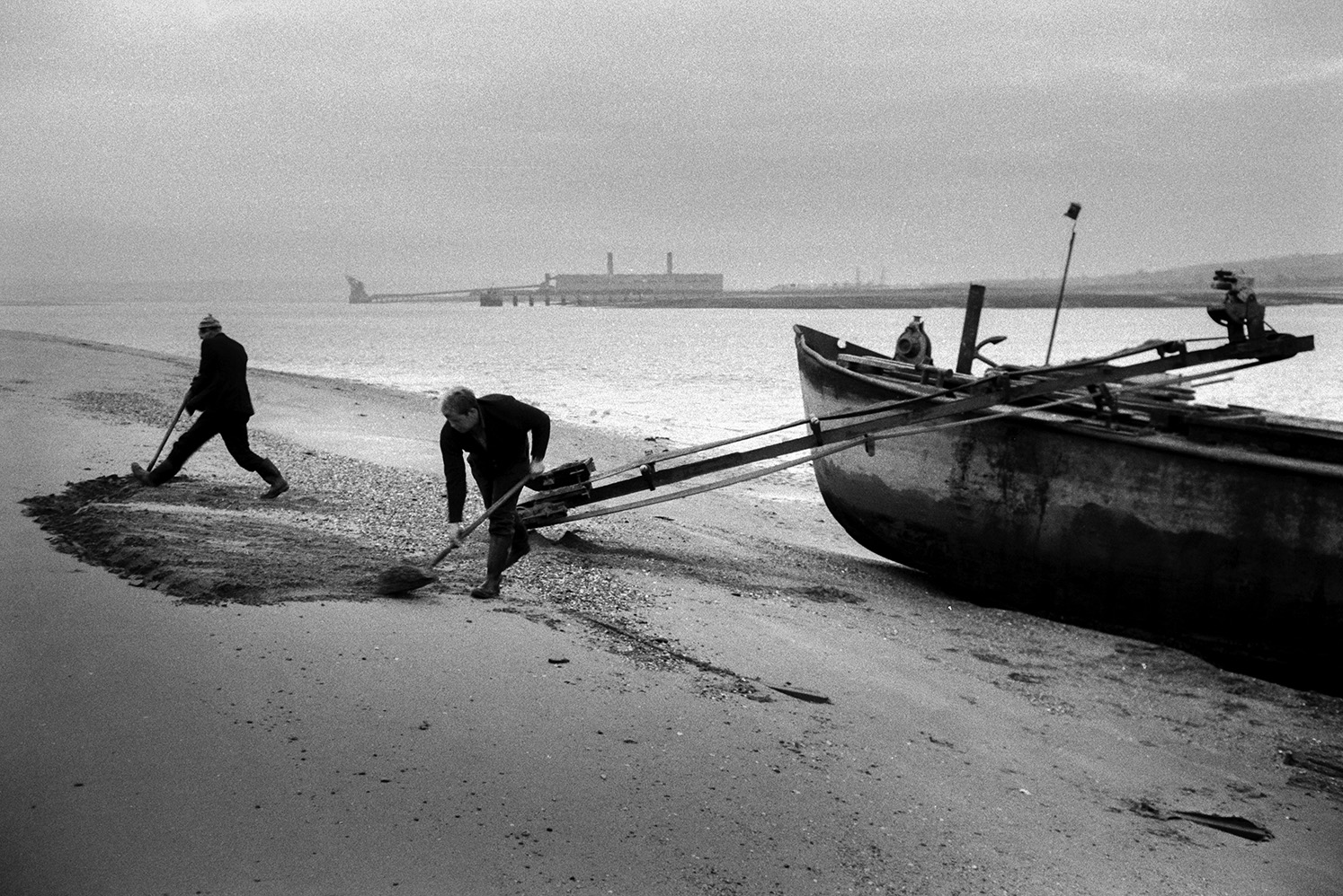 Men shovelling sand from the beach onto a conveyor belt or elevator attached to a sand barge at Crow Point, Braunton. The Taw River estuary can be seen in the background, as well as Yelland Power Station at Instow.