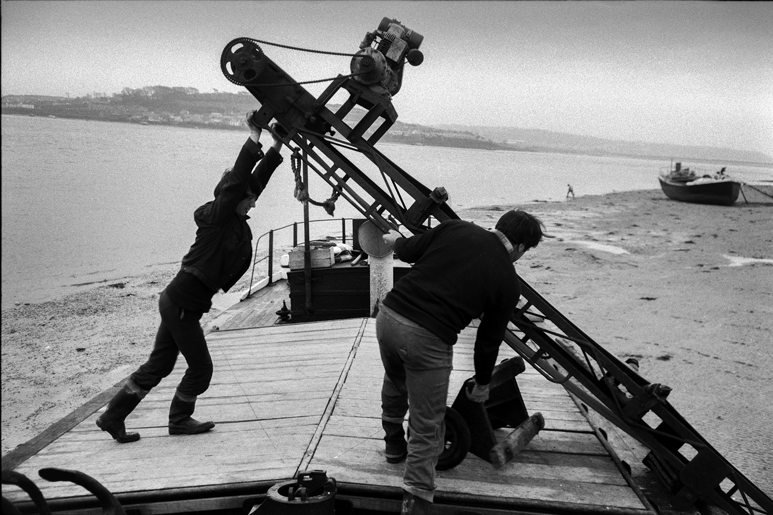 Men moving a conveyor belt or elevator off a sand barge and onto the beach at Crow Point, Braunton. The estuary of the Taw River can be seen in the background.