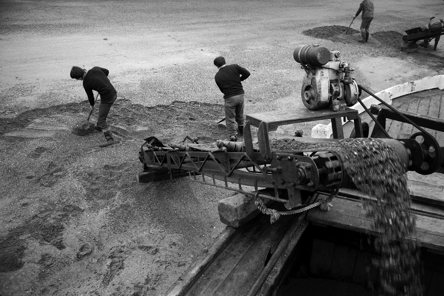 Men shovelling sand from the beach onto a conveyor belt or elevator attached to a sand barge at Crow Point, Braunton. The sand can be seen dropping into the barge at the top of the elevator.