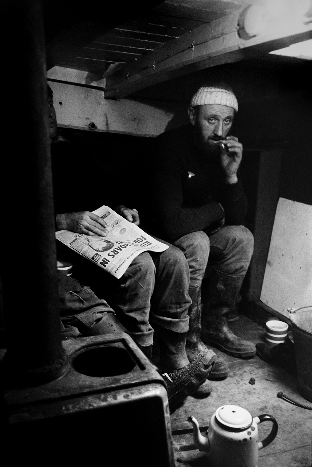 Men smoking and reading a newspaper inside a sand barge at Crow Point, Braunton. A teapot and mug can be seen on the floor.