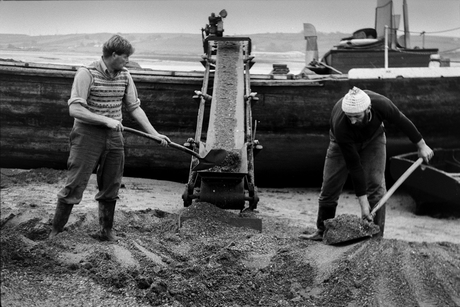 Men shovelling sand from the beach onto a conveyor belt or elevator attached to a sand barge at Crow Point, Braunton. The barge can be seen behind them.