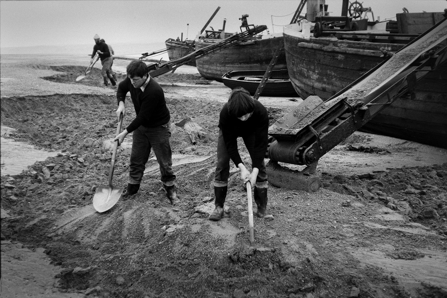 Men shovelling sand from the beach onto a conveyor belt or elevator attached to a sand barge at Crow Point, Braunton. The man on the left may be John Daniel. Two other barges being loaded with sand and a rowing boat can be seen behind them.