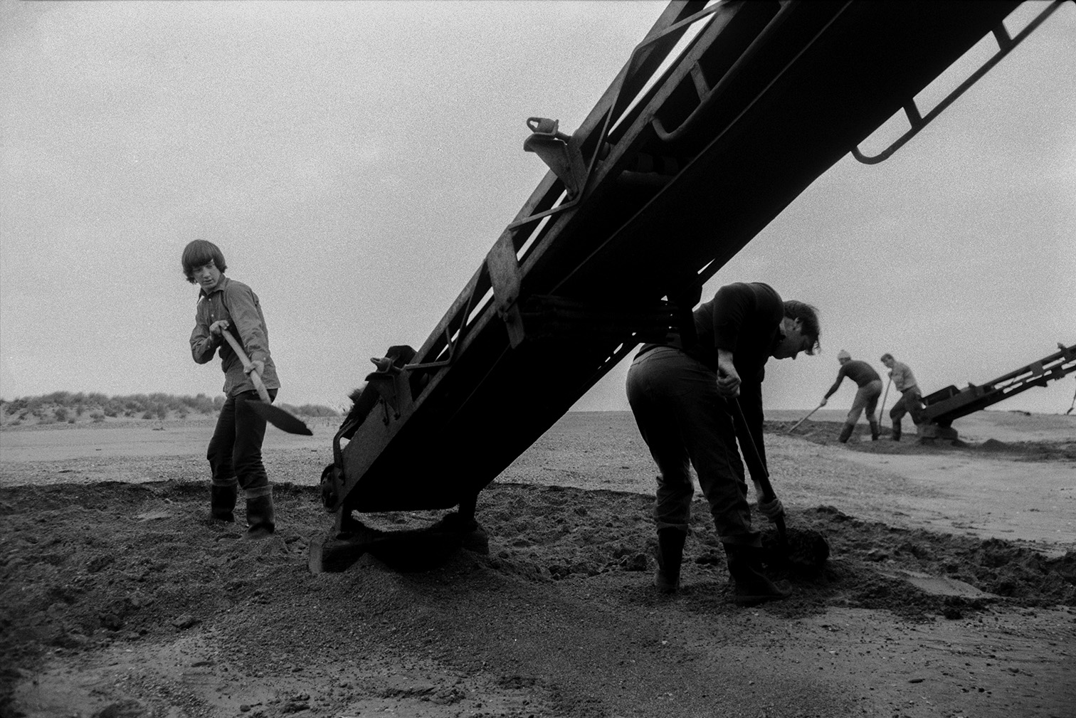 Men shovelling sand from the beach onto a conveyor belt or elevator attached to a sand barge at Crow Point, Braunton. Two other men loading a sand barge can be seen in the background.