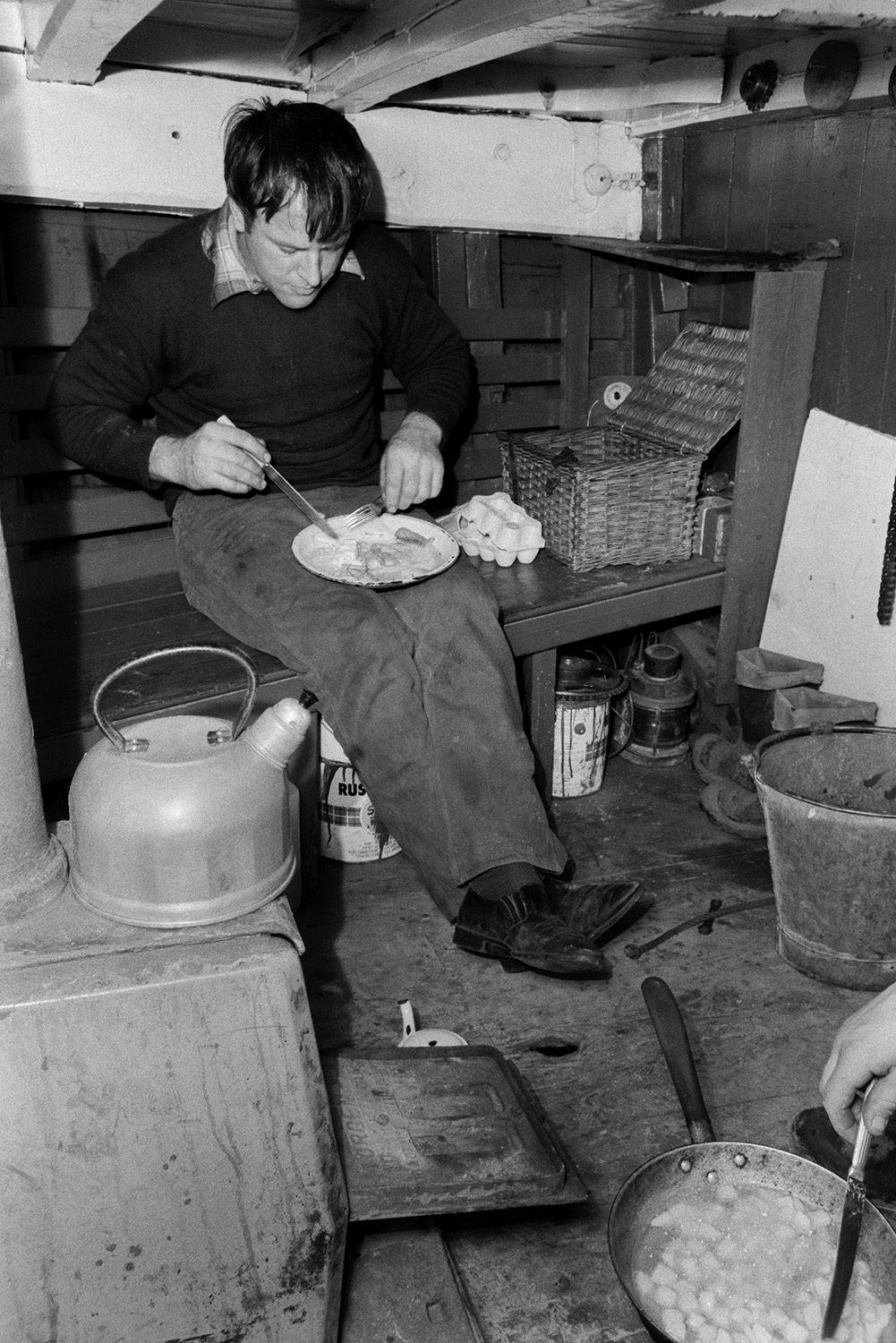 A man, possibly John Daniel, eating a fried breakfast inside a sand barge at Crow Point, Braunton, after loading the barge with sand from the beach. A basket, egg box, kettle and frying pan can be seen in the barge.