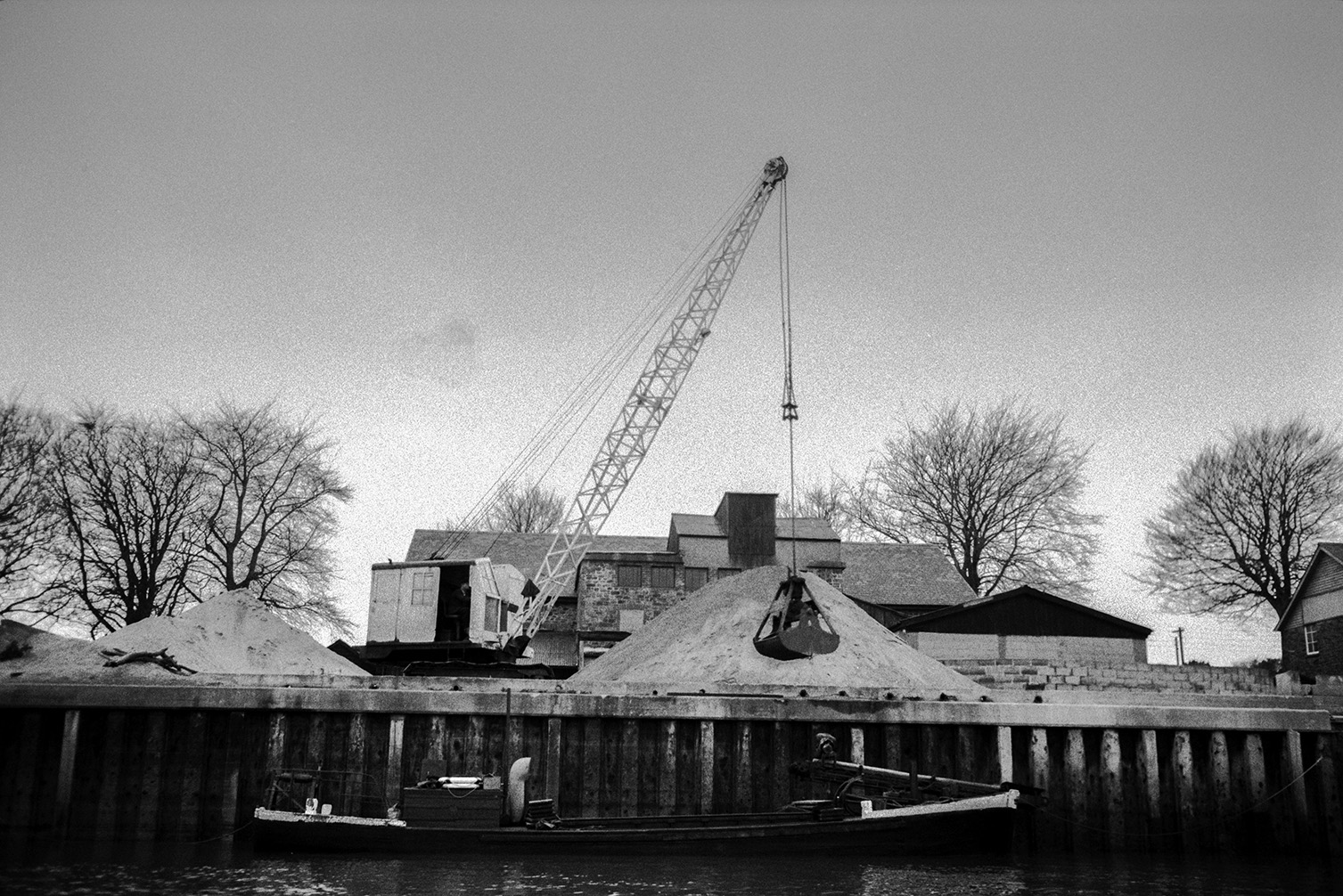 A crane with a mechanical grab unloading sand from a barge onto a dockside, possibly on the Taw River estuary.