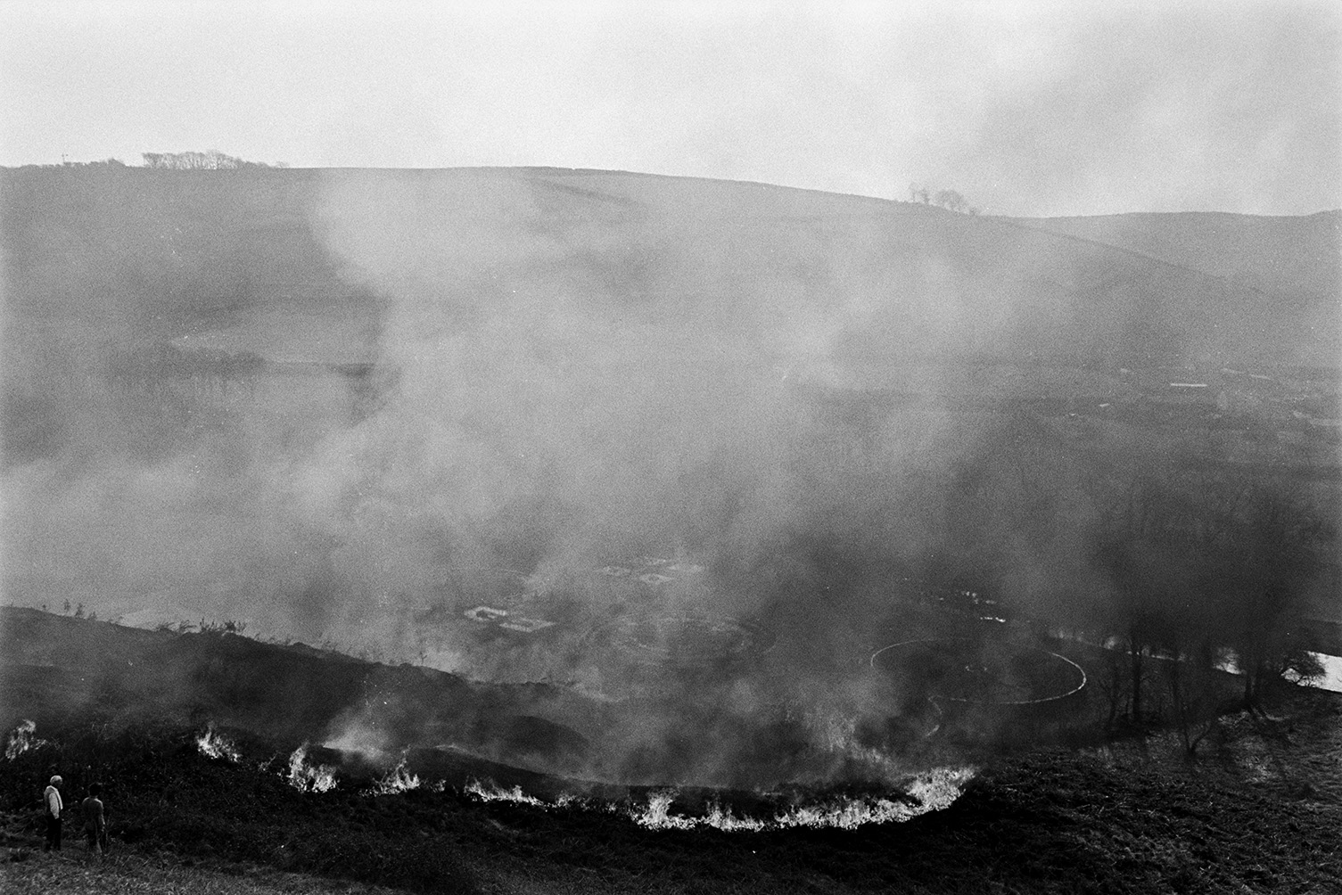 A fire on Torrington Hill to burn of surface brambles. Two people are watching the fire in the foreground. The River Torridge and a landscape of fields is visible in the background, through the smoke.