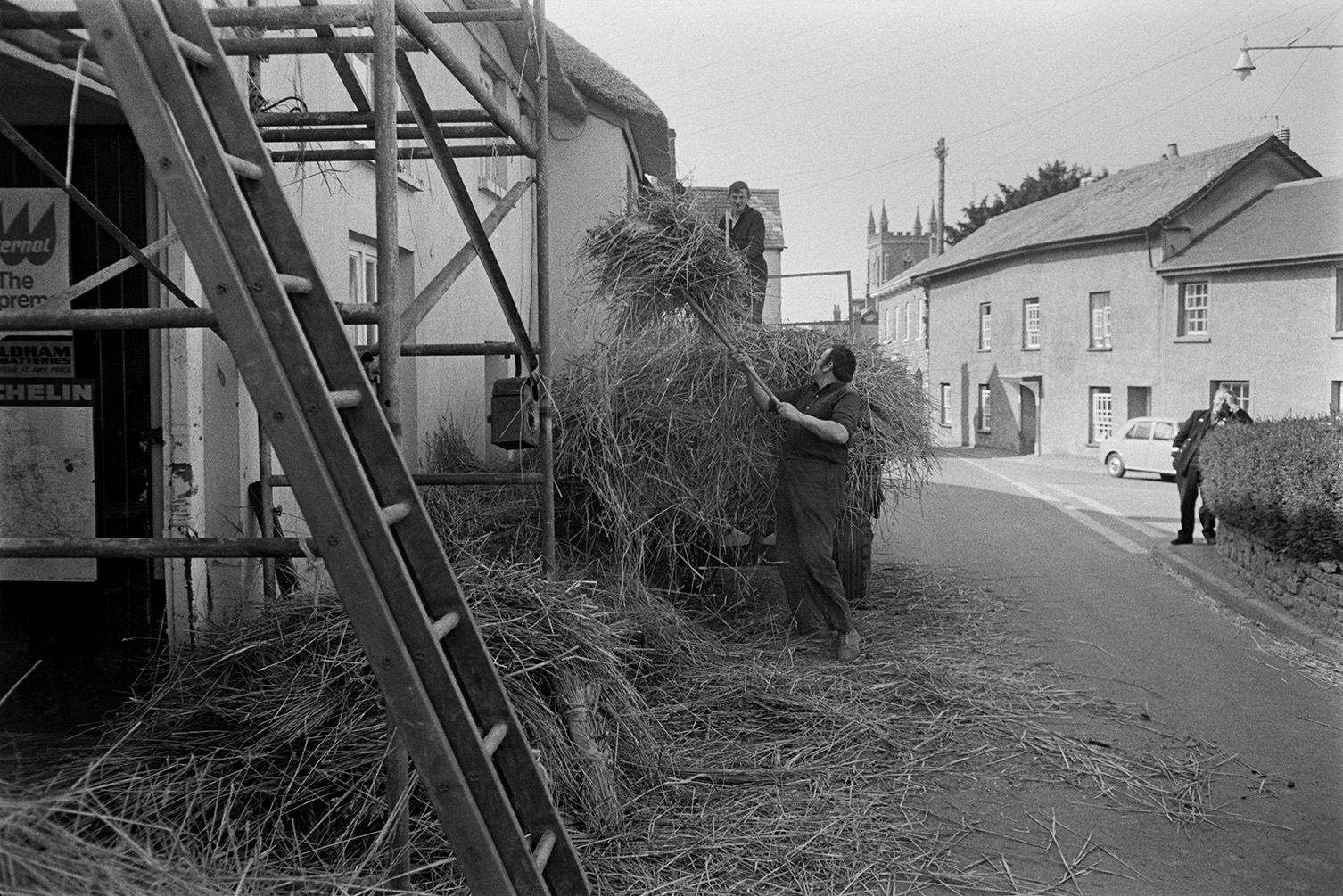 Two men loading old thatch reed which has been taken off a roof, onto a tractor and trailer in a street in Witheridge. They are using pitchforks. Scaffolding and a ladder are by the building being thatched. A man is watching from the other side of the street and Witheridge Church tower can be seen in the background.