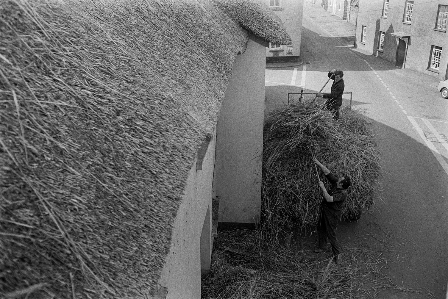 View of men loading old thatch reed onto a trailer in a street in Witheridge. The image is  taken from the roof top which is being thatched.