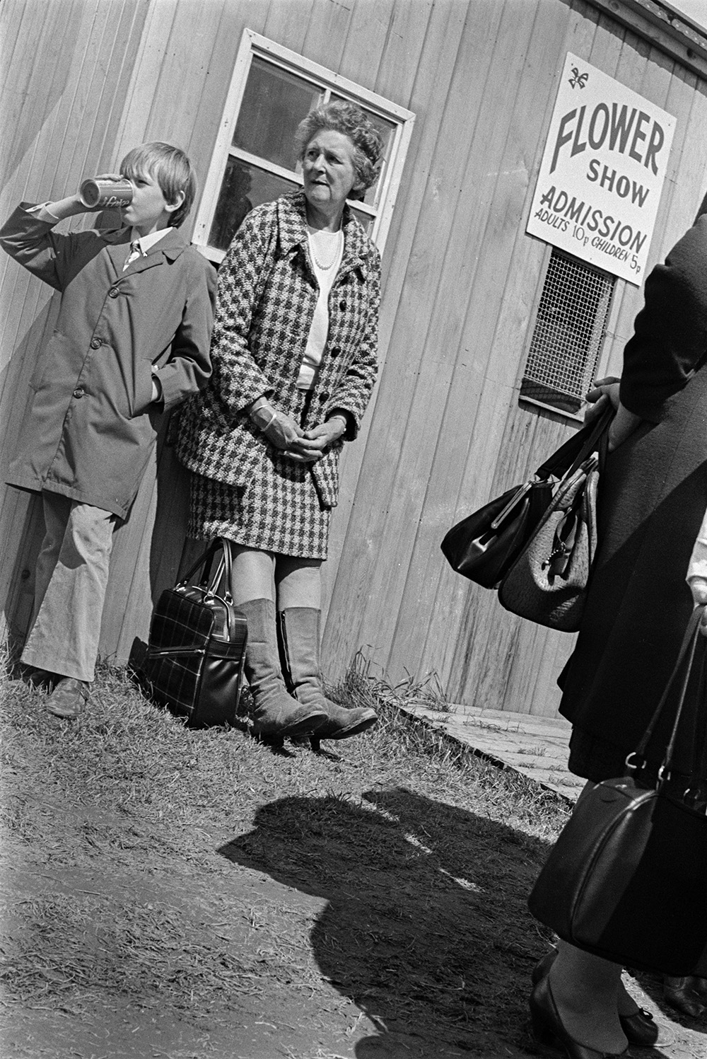 A woman and boy visiting the Devon County Show at Whipton, Exeter. They are stood outside the flower show admission hut. The boy is drinking from a can.