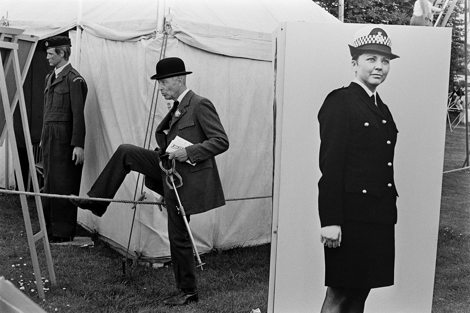 A man wearing a bowler hat climbing over a rope barrier by a show tent at the Devon County Show in Whipton, Exeter. A mannequin wearing a military uniform is in the background and a large photograph of a policewoman is on a display board in the foreground.