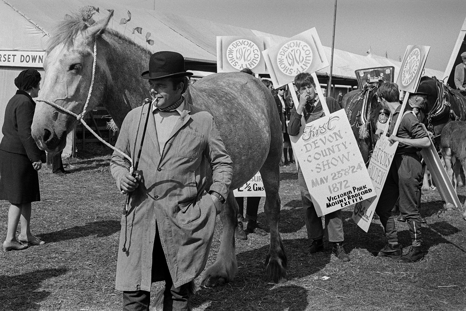 A Horse being led through visitors at the Devon County Show in Whipton, Exeter, by a man wearing a bowler hat. Boys in the background re wearing sandwich boards advertising the Show.