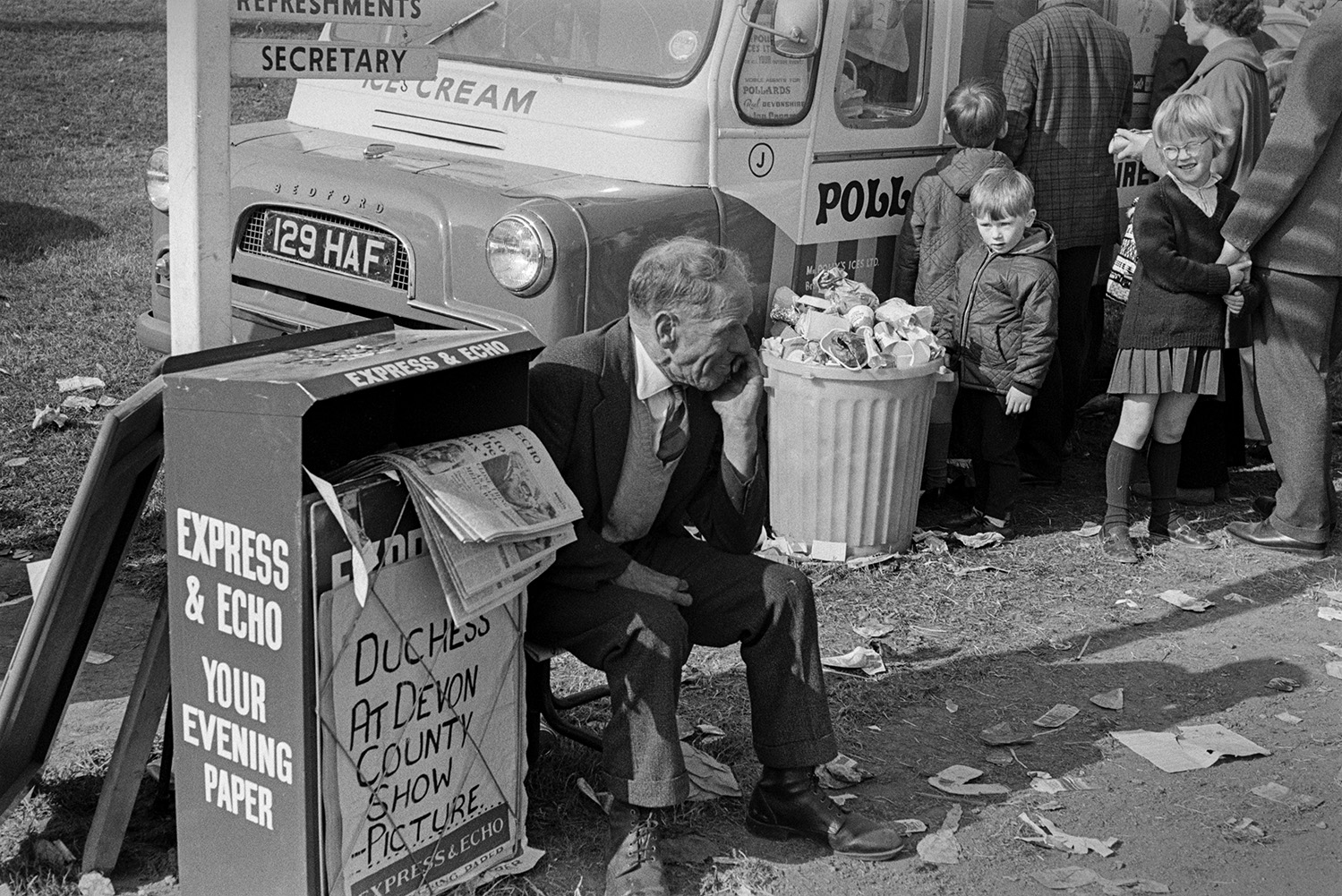 A newspaper vendor selling the Express and Echo newspaper at the Devon County Show in Whipton, Exeter. Men, women and children are queuing at a ice cream van, and rubbish is strewn on the ground from a overflowing bin, in the background.