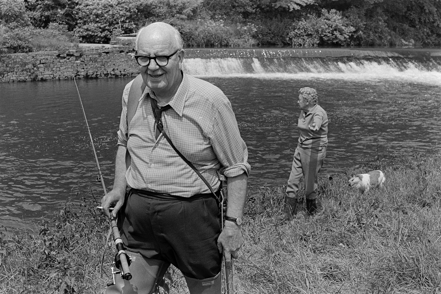 Dr J V Morris, OBE, by the River Torridge on a fishing holiday at the Half Moon Inn, Sheepwash. He is wearing waders and carrying a fishing rod. A woman and a dog are walking behind him along the riverbank and a weir can be seen in the background.